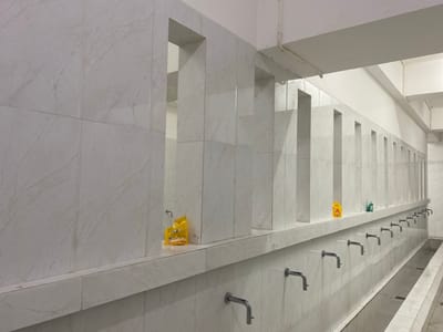 The image shows a row of sinks with faucets in a room that appears to be a public restroom or ablution area. The walls are tiled with a white marble pattern. There are several openings in the wall above the sinks, creating cubbies or stalls. On the shelf above the sinks, there are two small packets of what appears to be soap or cleaning product. The faucets are made of metal and are positioned along the wall. In the foreground, a long, shallow drain runs along the floor. The overall impression is of a clean but utilitarian space. The scene is indoors and appears to be a well-lit area, though the light source is not visible. No people are present in the image.