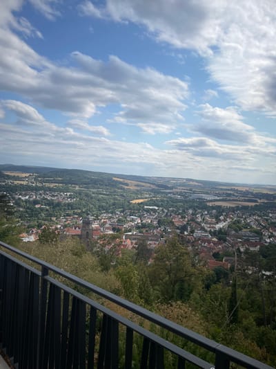 The image is an aerial view of the city of Homberg (Efze), Germany, taken from a high vantage point. A black metal railing in the foreground suggests the photo was taken from a balcony or viewing platform. Below the railing, a dense green forest slopes down towards the city. The city itself is characterized by red-tiled roofs and a prominent church tower with a dark, weathered facade. The urban landscape is nestled within a valley, with rolling hills and agricultural fields stretching into the distance under a sky filled with scattered white clouds. The overall impression is of a picturesque, historic European town surrounded by nature.