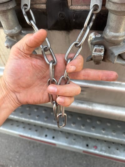 A person's hand and fingers are shown in a close-up shot, gripping a metal chain. The chain is attached to a larger metal object in the background, which appears to be part of a vehicle or industrial equipment. The background also shows a textured metal surface with numerous circular indentations, suggestive of a ramp or a walkway. The ground beneath is visible and appears to be asphalt. The lighting suggests it is daytime. No specific location in the city of Lebanon, United States of America, can be identified from the visual information provided.