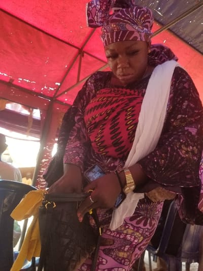 The image depicts a woman in a semi-enclosed setting in Ochadam, Nigeria. She has a dark complexion and wears a multi-patterned headwrap and a loose-fitting garment, both featuring designs in purple, red, black, and yellow. A white fabric is draped over her left shoulder. She is observed holding a black smartphone displaying "1:40" in her left hand, which also wears a gold-toned watch and a silver-colored ring. Her gaze is directed downwards as she interacts with a black, textured handbag, possibly opening or searching its contents. The immediate background shows fragments of blue plastic chairs. Overhead, a red fabric canopy structure is visible. A blurry figure is partially discernible in the distant left background.