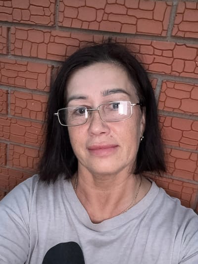 The image is a selfie of a middle-aged woman with short, dark hair, wearing glasses and a light-colored t-shirt. She is standing in front of a brick wall. The bricks are reddish-brown with dark grout lines. The lighting suggests it is daytime. The woman is looking directly at the camera with a neutral expression. There is a thin gold chain necklace visible around her neck. The image does not contain any information or visual cues that would allow for the determination of a specific location within the city of Malmyzh, Russia.