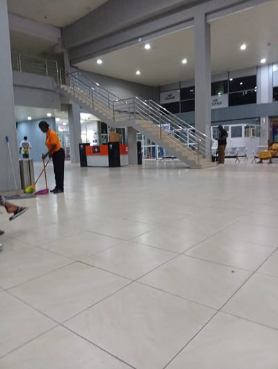 The image depicts an interior view of Port Harcourt International Airport in Omagwa, Nigeria. The setting is a large, open hall with tiled flooring. In the foreground, a woman in an orange shirt and black pants is sweeping the floor with a broom. Nearby, a metal trash bin and a mop are visible. A staircase with metal railings leads to a higher level where signs indicating a "VIP Lounge" are displayed. In the background, individuals are seated on chairs. A service counter is visible behind the cleaning woman. The overall environment suggests a public waiting area within the airport.