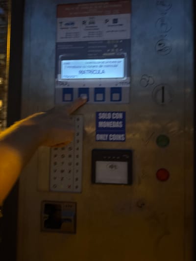The image shows a close-up of a parking payment machine in Calp, Spain, likely outdoors. A person's hand is visible on the right side of the frame, with their index finger pressing one of the buttons on the machine. The machine has a digital display showing "MATRICULA" and "Introduzca el número de matrícula" which translates to "Enter the license plate number." There are also various buttons, slots for coins, and what appears to be a contactless payment reader. The machine itself is metal and shows signs of wear and tear, with some graffiti or markings on its surface. The background is blurred, but suggests an urban environment, possibly a street or parking lot. The lighting suggests it might be dusk or nighttime.