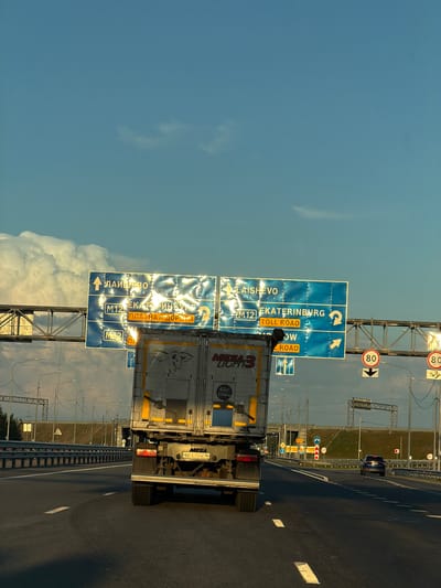 A truck travels down a highway in Egor'evo, Russia, under a clear blue sky with large, white clouds. The truck, with a graphic of a bull on its side, is positioned in the right lane, moving away from the viewer. Overhead signs indicate directions to "LAISHEVO" and "EKATERINBURG" with "TOLL ROAD" highlighted in orange. The highway has multiple lanes and is bordered by a metal guardrail and lampposts. In the distance, other vehicles, including a dark SUV, can be seen on the road. Speed limit signs showing "80" are visible on the right side of the highway. The sun appears to be setting, casting a warm light on the scene.
