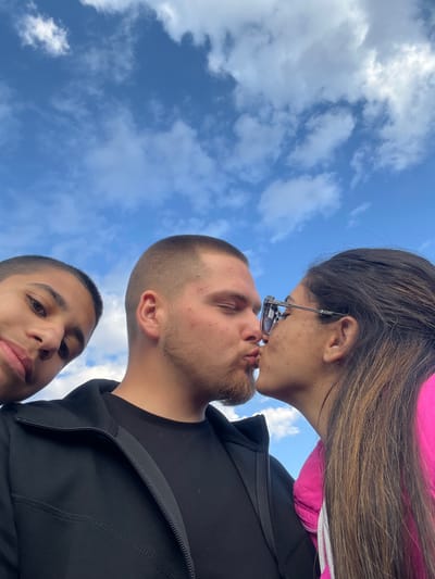 A low-angle photograph shows three people against a sky with scattered clouds. A woman with long brown hair and glasses is kissing a man with a short haircut and beard. A younger person with short hair is visible at the bottom of the frame. The man wears a black jacket, and the woman wears a pink shirt beneath her black jacket. The location is unknown and there is no indication of Kribul, Bulgaria.