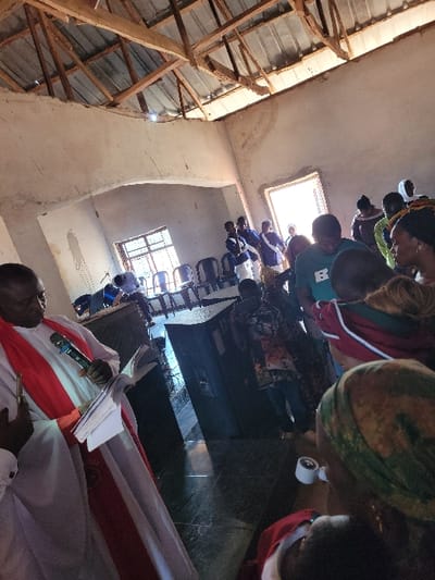 The image depicts an indoor gathering within a functional building in Rinze, Nigeria. The structure features light-colored, visibly worn walls with an arched opening, and an exposed roof comprising wooden rafters and corrugated metal sheeting. Multiple unglazed window openings admit bright natural light, illuminating a dark, reflective floor.
In the foreground left, a dark-skinned male, attired in a white robe with a red stole, holds a blue and silver microphone in his right hand and papers in his left, facing the assembled individuals. To his right, two large black speaker enclosures are positioned on the floor. The room is populated by numerous people. A group of young males in blue and white uniforms stands in the mid-background near several stacked black plastic chairs. Other attendees, including a woman wearing a patterned green headscarf and holding a white mug in the foreground right, are present in various civilian clothing and head coverings. A child's head is partially visible in the immediate foreground right. The overall scene indicates a community or religious event in progress.