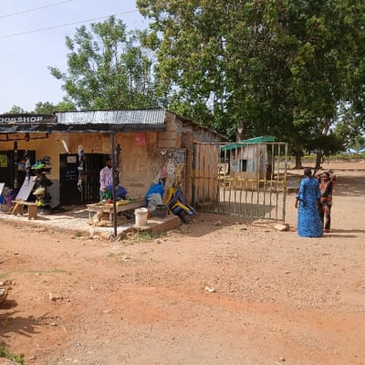 The image shows a small, unpaved street in Kufang, Nigeria, located at coordinates 9.863 latitude and 8.867 longitude. A single-story, concrete building with a corrugated metal roof stands on the left side of the street. The building is labeled "BOOKSHOP," and its exterior is painted a pale yellow. There are several shoes displayed on the shop's exterior wall. A man wearing a red and white patterned shirt and dark pants stands just outside the shop's entrance. A small, wooden bench with a white tabletop sits in front of the shop. There are several other objects visible on the ground near the bench, including a large blue plastic tub, a large, blue plastic container, and a variety of assorted, colorful plastic chairs. There is a metal gate that closes off the street to vehicular traffic at the entrance. Two individuals, a man and a woman, are walking through the gate. The woman is wearing a long, blue patterned dress, and the man is wearing a white shirt and blue pants. Behind the gate, on the right side of the street, there is a small, concrete building, painted green and yellow. It is a simple, rectangular structure with a corrugated metal roof. The background is a dirt field covered with green vegetation. There are large, mature trees in the background. The sky is a clear, light blue. The image appears to have been taken on a sunny day, and the weather is warm and dry.
