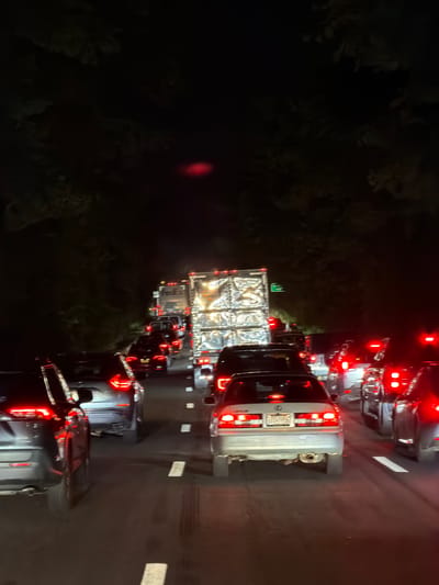 The image depicts a nighttime traffic jam on a highway in the Village of North Hills, United States of America. The road is packed with numerous vehicles, including cars and at least two large trucks, one of which is branded with "FedEx". The taillights of the vehicles are illuminated, casting a red glow on the asphalt. The surrounding environment appears to be dark, with trees lining the edges of the highway. The overall scene suggests a significant delay and congestion on the roadway.