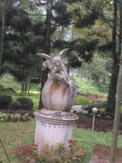 A weathered stone statue is centrally positioned in an outdoor garden environment in Ede, Netherlands. The statue depicts a light-colored, winged figure with prominent wings and a human-like torso, posed in a seated or reclining position atop a spherical element. The figure's left hand is near its chin. This sphere rests on a fluted, cylindrical pedestal. Both the statue and pedestal exhibit significant discoloration, weathering, and dark staining. The immediate foreground consists of green grass and patches of reddish-purple and green foliage. The midground features landscaped garden beds with manicured dark green bushes, rows of reddish-purple plants, and green ground cover. A light-colored spherical garden lamp on a thin pole is visible to the right of the pedestal. A narrow, curving path extends into the distance on the right. The background is composed of numerous tall, dark green coniferous trees with dense foliage, interspersed with lighter green deciduous trees further back. The scene is illuminated by diffused natural light, consistent with an overcast sky. No individuals are present.