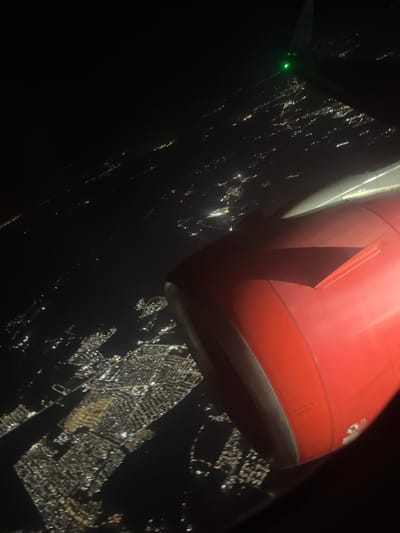 An aerial view at night shows a sprawling city grid illuminated by countless lights. A large, red airplane engine dominates the upper portion of the frame, positioned above the city. A part of a wing with a green light can be spotted in the upper left corner. The scene is dark, emphasizing the contrast between the darkness and the glowing city below.