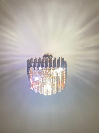 A wall-mounted light fixture dominates the frame. The fixture features a circular design with horizontal metal slats and rows of clear, rectangular crystals that emit a bright glow. Light rays radiate outward from the fixture, creating shadows on the surrounding off-white wall. The wall is softly illuminated by the emitted light.