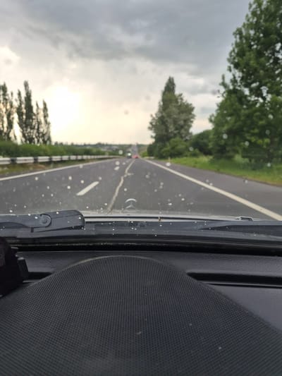 The image shows a view from inside a car driving on a highway, likely in the vicinity of Hrazdan, Armenia, under a cloudy sky. The foreground is dominated by the car's dashboard and windshield wiper. The windshield has water droplets on it, indicating recent rain or dew. The highway stretches into the distance, with a white line marking the lanes. Trees and greenery line the sides of the road, and a guardrail is visible on the left. In the far distance, the road appears to curve, and distant lights suggest a town or city. The overall atmosphere is overcast and somewhat gloomy. There are no people visible in the image.