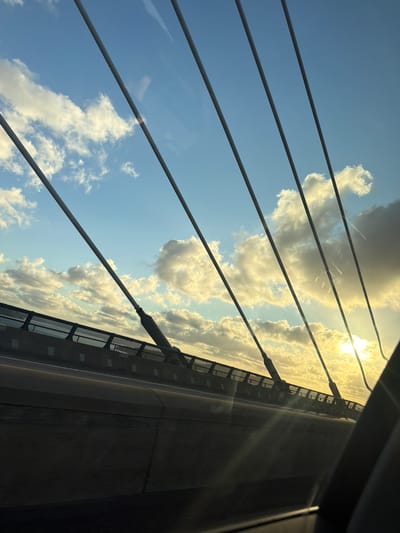 The image captures a view from a vehicle traveling on a bridge, likely a cable-stayed bridge given the prominent suspension cables visible. The scene is dominated by a bright sunset with the sun partially obscured by clouds, casting a warm glow across the sky and illuminating the clouds from behind. The sky is a vibrant blue with scattered white and gray clouds.

In the foreground, the interior of the vehicle is visible on the right side, with a dark pillar obscuring part of the view. The bridge's structure is evident, with a roadway and a barrier along the edge. The suspension cables, which are dark gray, run diagonally across the frame, connecting to the bridge deck.

The location is specified as Ödsmål, Sweden, suggesting this bridge is a significant piece of infrastructure in that area. No people are visible in the image. The overall impression is one of a scenic journey during sunset, highlighting the architectural elements of the bridge against a dramatic natural backdrop.