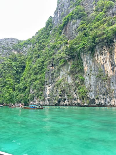 The image depicts a scenic view of limestone cliffs covered in lush green vegetation, rising dramatically from the turquoise waters of Haad Yao, Thailand. Several longtail boats are anchored near the shore, with some carrying passengers and others appearing empty. The water is clear and calm, reflecting the surrounding cliffs and the overcast sky. The cliffs are characterized by their rugged, rocky surfaces and the dense foliage that clings to their steep slopes. The overall scene conveys a sense of natural beauty and tranquility, typical of tropical island landscapes.