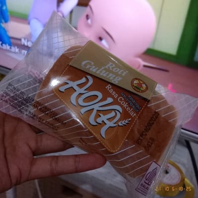 A package of "Roti Gulung" (rolled bread) with chocolate flavor is displayed in front of a cartoon image on a screen. The brand name "AOKA" is prominently featured. The packaging indicates the product is a variant of chocolate flavor, produced by PT. Indonesia Bakery Family, and Halal certified. The label includes the BPOM RI MD number and the expiration date: EXP04092025, 0418. The time stamp "21:10 6-18-25" is visible in the upper right corner. A hand is holding the product.