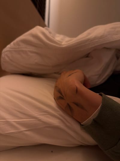 Within a location identified as Kutchan, Japan, an indoor scene is depicted under warm, dim artificial lighting. The primary subject is a human right arm and hand, resting horizontally on a white pillow and white bed sheet. The forearm is covered by a muted olive-green long-sleeved garment with a ribbed white cuff. The back of the hand and wrist feature a dark, temporary marking resembling a radial pattern of multiple leaf-like or feather-like shapes, with curving lines extending towards the wrist and an incomplete circular outline. Some of the markings appear slightly smudged. A crumpled white duvet or sheet is visible in the upper portion of the frame, partially covering the bedding. The background is blurred, revealing a light-colored wall and a dark, gridded fixture in the upper left, consistent with an interior room. No active movement or interaction is observed.