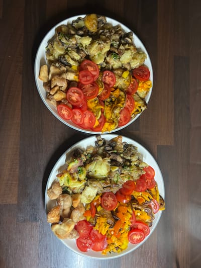 Two white plates filled with a colorful mixture of food are placed on a dark wooden surface. Each plate contains roasted potatoes, mushrooms, chopped chicken pieces, sliced tomatoes, and what appears to be bell peppers or carrots. The lighting suggests an indoor setting, possibly a home kitchen or dining area. There is no indication of the city of Balbriggan, Ireland, or any specific urban environment.