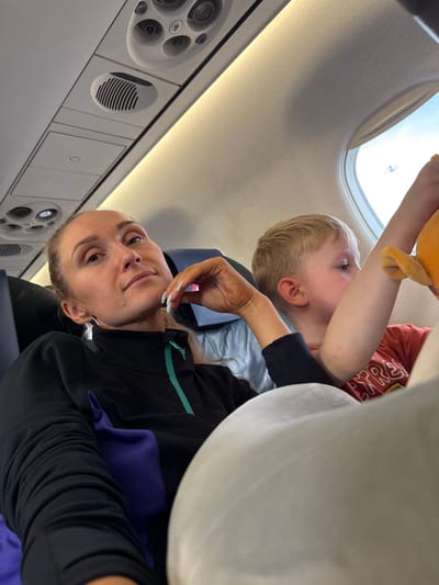 A woman and a young boy are seated next to each other on an airplane. The woman, in the foreground, is looking towards the camera with her head tilted and her hand resting on her chin. She is wearing a black zip-up jacket with a teal accent. The boy, in the background, is facing away from the camera, looking out the airplane window. He is wearing a red t-shirt and holding a stuffed toy. The interior of the airplane cabin is visible, including overhead compartments, air vents, and the window. The image is taken from a slightly elevated angle. There is no discernible indication of the city of Bol'shie Kabany, Russia in the image.