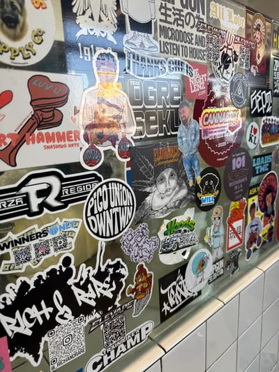The image shows a close-up of a wall covered in stickers. The stickers feature various designs, including cartoon characters, text, and logos. Some of the text is in English, while other text appears to be in Japanese. The wall itself is a metallic surface, and it is partially covered by white tiled surfaces at the bottom. There are no people visible in the image. The overall impression is that of a decorated wall, likely in an establishment or public space. The location is specified as being in Culver City, United States of America.