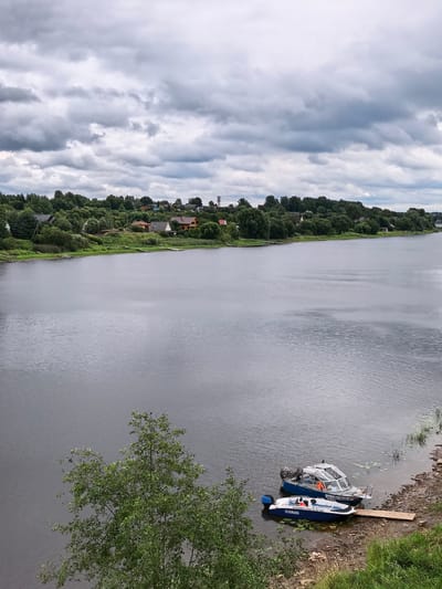 An elevated view of a wide, dark river flows towards the viewer under a heavily overcast sky. On the distant shore, a line of dense green trees forms a backdrop for scattered houses and buildings, with a water tower visible on the far right. In the foreground, the river's surface is calm, with ripples suggesting a gentle current. Two boats are moored at the edge of the water, resting on a wooden pier that extends into the river. The larger boat is blue and silver with a covered cabin and an outboard motor, and bears the registration "AX 0365 RUS 47". A smaller blue and white boat, a Yamaha, is positioned in front of it. A green tree with lush foliage occupies the lower left corner of the frame, partially obscuring the river and shoreline. The scene appears to be in Staraya Ladoga, Russia.