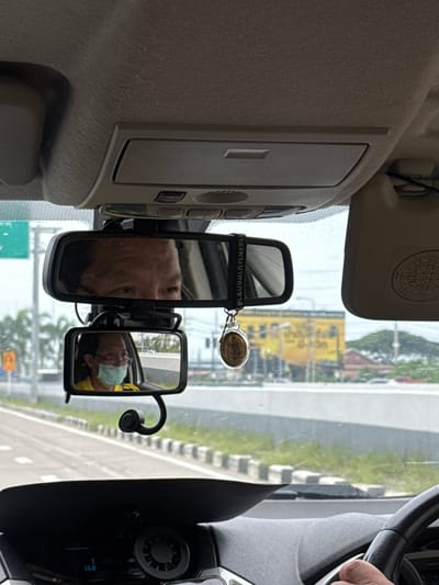 The image shows the interior of a car, as viewed from the driver's perspective. The driver's face is reflected in the rear-view mirror, showing them wearing glasses and a light blue face mask. A secondary, smaller mirror attached below the main rear-view mirror also reflects the driver, along with the interior of the car and the road ahead. The dashboard and steering wheel are visible, with a digital display showing "14.0". A coin amulet hangs from the rear-view mirror. Outside the car, a highway is visible with a yellow building and trees in the background. The location is identified as Tha Sala, Thailand, based on context not present in the image itself. The scene captures a moment of driving, with the driver's focus likely on the road.