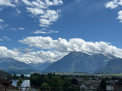 The image shows a panoramic view of the city of Thun, Switzerland, with the Alps in the background. The foreground features a body of water, likely Lake Thun, with several boats docked along its edge. Dense green trees line the lake, and beyond them, a town with a mix of residential and commercial buildings is visible. The distant mountains are majestic, with snow-capped peaks contrasting against a bright blue sky dotted with fluffy white clouds. The overall scene depicts a serene and picturesque landscape characteristic of the Swiss Alps.