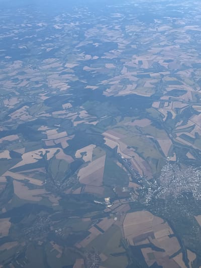 This aerial photograph captures a vast, cultivated landscape viewed from a high altitude. The terrain is a mosaic of agricultural fields, characterized by irregular shapes and varying shades of brown, beige, and green, indicating different crops or stages of growth. Patches of dark green denote forested areas, interspersed with the fields. A small town, identified as Novo Panicharevo, Bulgaria, is visible on the right side of the image, appearing as a dense cluster of buildings with a grid-like street pattern. The town is surrounded by the agricultural land and stretches towards the lower right corner of the frame. The overall impression is one of rural expanse with a concentrated settlement.