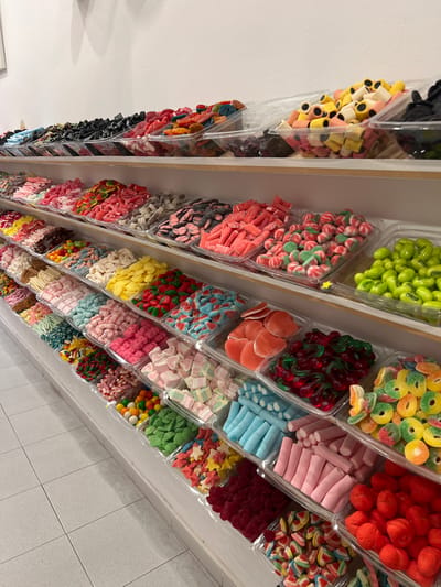 The image shows a display of bulk candies in clear plastic containers, arranged on white shelves against a white wall. The candies are varied in color, shape, and type, including licorice, gummy candies, marshmallows, and fruit-flavored candies. The display is likely in a candy shop. The floor is tiled in a light gray color. The location is the city of Port de Pollença, Spain.
