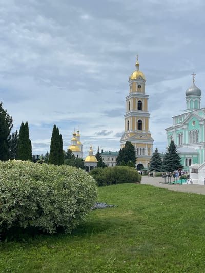 The image depicts a scenic view of the Seraphim-Diveyevo Monastery in Diveyevo, Russia. In the foreground, a lush green lawn with scattered small white flowers stretches across the frame, partially obscured by a large, variegated green and white bush on the left. The bush has a dense canopy and appears well-maintained. Behind the bush, a portion of a dark-colored object, possibly a tarp or bag, lies on the grass.
Dominating the mid-ground and background are several prominent architectural structures of the monastery. A tall, yellow bell tower with a golden dome stands prominently, featuring multiple levels with arched windows and a clock face. To its right is a building with a light blue facade and a silver dome, adorned with white decorative elements. Further to the left, several golden-domed churches are visible, some with white walls and intricate architectural details. Tall, dark green evergreen trees are strategically placed throughout the scene, adding height and contrast to the landscape.
A paved walkway or courtyard is visible in the mid-ground, where several people are scattered. Some are standing, others are seated on benches. They appear to be casually strolling or observing the surroundings. The sky above is overcast with a mix of gray and white clouds, suggesting a slightly diffused lighting condition. The overall atmosphere of the image is serene and picturesque, capturing the grandeur of the monastery and its peaceful grounds.