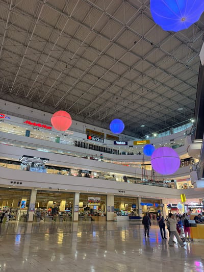The image shows the interior of a multi-level shopping mall, likely in the city of Tekirdağ, Turkey, given the presence of Turkish signage such as "MediaMarkt," "Mudo," "Evidea," "Vatan," "Cepax," and "Carrefour." The ceiling is a complex metal truss structure. Large, illuminated spherical decorations in red, blue, and purple are suspended from the ceiling. Several floors of shops are visible, with prominent store names displayed on the upper levels. On the ground floor, a Carrefour supermarket is open, with checkout counters visible. Several people are walking through the mall, with some gathered near shops and others in the foreground. The floor is polished and reflects the lights and store displays. The overall atmosphere appears to be that of a busy commercial center.