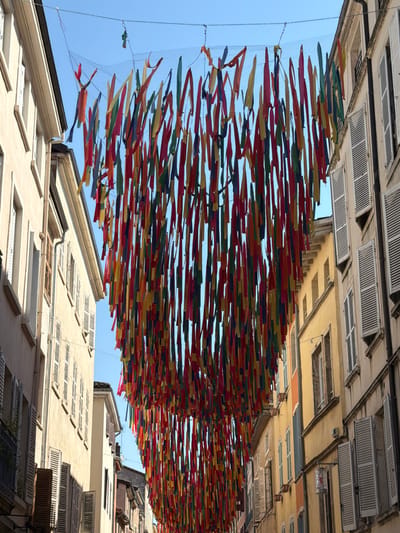 In Charnay-lès-Mâcon, France, a street is adorned with a vibrant canopy of colorful streamers suspended between buildings. The streamers, in hues of red, yellow, blue, green, and orange, hang from a mesh net stretched across the street, creating a festive and dynamic visual. The buildings lining the street feature traditional architecture with shuttered windows and a mix of cream and yellow facades. The sky above is clear and blue. The scene suggests a celebratory atmosphere or an event taking place in the town.