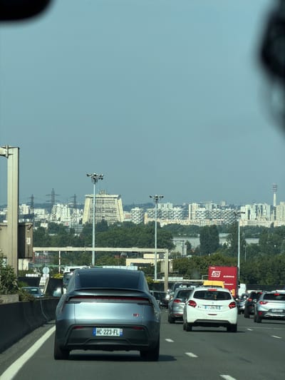 The image captures a view from inside a vehicle traveling on a highway. In the foreground, the rear of a light grey electric vehicle with license plate HC-223-FL is visible. It is in the right lane of a multi-lane highway. Beyond this vehicle, several other cars are in traffic, including a white Peugeot 208 with license plate FJ-294-DL, a silver sedan, and other vehicles. A large red truck with "SD SLOVAKIA" and "TRANSPORT" branding is also present in the traffic.
In the background, a cityscape is visible under a pale blue sky. Prominent structures include several tall buildings with a unique, tiered architectural style, possibly apartment complexes. In the mid-distance, there is a distinctive, angular, pyramidal-shaped building with numerous windows. Power lines and towers are also visible, indicating infrastructure within the urban environment. The overall scene depicts a traffic jam on a highway with an urban backdrop, suggesting a location within or near a populated area. The presence of these landmarks suggests the image was taken on the A86 highway in Thiais, France.