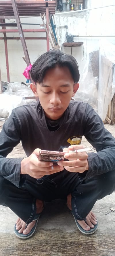 A young male is depicted squatting on a light grey, worn concrete surface in Cilampeni, Indonesia. He has dark hair, closed eyes, and a neutral facial expression. He wears a dark grey long-sleeved shirt with a light yellow graphic of a cup, dark pants with visible white stitching, and dark blue flip-flops. His left hand holds a smartphone encased in a reddish-brown cover featuring a character graphic. His right hand holds a lit cigarette with visible ash between his index and middle fingers, positioned above the smartphone. A metal ring is attached to the left side of his pants.

The background consists of a light-colored wall with visible pipes and two wooden shelves. The upper shelf holds a black bag, a spray bottle, and various containers. The lower shelf supports a black electric water pump with exposed wiring and a two-prong plug. To the left, a reddish-brown metal framework is visible, with a pink fabric item draped on it and white plastic sheeting or debris piled on the ground. A large, clear plastic tarp is draped vertically on the right side of the frame.
