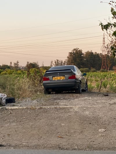 This image, captured in Sidi Moussa, Algeria, depicts a dark grey sedan car parked on an unpaved road during what appears to be dusk or dawn. The vehicle, a BMW, is viewed from its rear three-quarter angle, with its back facing away from the viewer. A yellow license plate is clearly visible on the trunk, displaying the characters "03972 195 16".

To the right of the car, the terrain is characterized by overgrown, wild vegetation, including green and brown shrubs, with a line of trees in the farther distance. A large metal electricity pylon stands prominent in the mid-ground, and multiple power lines stretch across the light, yellowish-orange sky.

On the immediate left side of the vehicle, a textured, light grey concrete or paved surface extends into the foreground. Near the car's rear bumper, on the ground, a black container or bin is present alongside some crumpled white material, possibly discarded plastic bags.

The scene conveys a quiet, semi-rural atmosphere, with no people visible and no specific activity taking place other than the car being stationary. The soft, warm light of the sky suggests either early morning or late afternoon, possibly with some atmospheric haze. The "16" on the license plate is a geographical code identifying the Algiers region in Algeria, aligning with the provided location context.