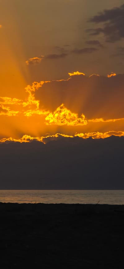 The image depicts a sunset over a body of water, likely a lake or sea, with a dark foreground. The sky is filled with dramatic clouds illuminated by the setting sun, creating rays of light that pierce through them. The clouds are a mix of bright orange and yellow near the sun, transitioning to darker shades as they move away. Below the sky, the water reflects the colors of the sunset, appearing as a band of muted orange and yellow. The foreground is a dark silhouette, suggesting land or a beach with some indistinct shapes that could be vegetation or debris. The scene conveys a peaceful and serene atmosphere. The provided location of Basbeshik, Kyrgyzstan, is a mountain lake, which is consistent with the imagery of a vast body of water and a dramatic sky.