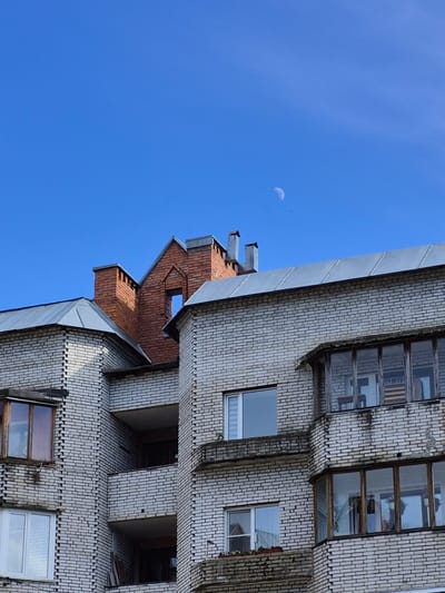 An outdoor, eye-level, medium shot captures the upper section of a light gray brick apartment building against a clear, bright blue sky. A crescent moon is visible in the upper right portion of the sky, accompanied by a small dark bird. The building features multiple windows, some with wooden frames and balconies, as well as brick chimneys on the roof. The architecture suggests a post-Soviet era style, common in many Russian cities. The lighting indicates it is daytime, possibly late afternoon given the angle of the sun. The image appears to be a detail of a residential building in the city of Pavlovo, Russia, though no specific landmarks or distinguishing features are visible that would pinpoint its exact location within the city. There are no people or animals present, other than the distant bird.