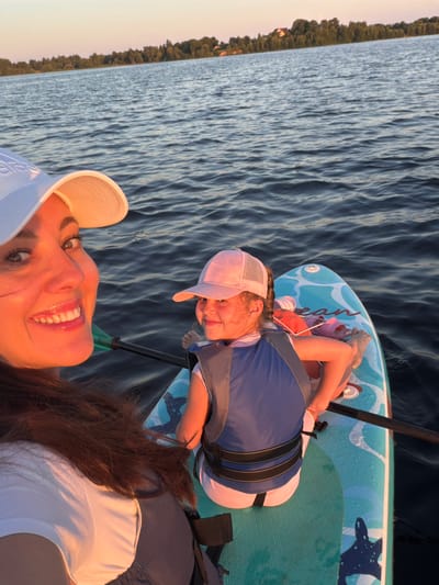 The image depicts a woman and a young girl on a paddleboard on a body of water, likely a lake or river, with a tree-lined shore in the background. The woman, in the foreground and to the left, is wearing a white baseball cap and a white t-shirt, with her right arm and shoulder visible. She is smiling and looking towards the camera. The girl is seated on the paddleboard behind her, wearing a pink baseball cap and a blue and grey life vest over a white and pink outfit. She is also smiling and looking towards the camera. The paddleboard is light blue with a white wave-like pattern and images of sea creatures. A paddle is visible extending across the board between the two individuals. The sun appears to be setting, casting a warm glow on the scene. The location is identified as Salaspils, Latvia.