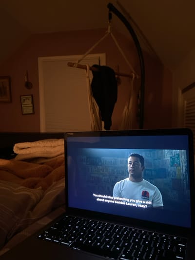 A laptop computer is open on a bed with a blanket, showing a man on the screen who is speaking. The laptop screen is showing a scene from a movie with a caption that reads, "You should stop pretending you give a shit about anyone besides Lauren, okay?" The laptop is positioned in front of a bed with a blanket on top of it. There is a hanging hammock in the background. The scene is in a bedroom located at 40.952, -73.708 in Rye, New York.