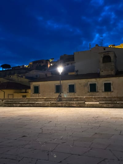 The image shows a night scene in Portoferraio, Italy, a town on the island of Elba, located at the coordinates latitude 42.816 and longitude 10.331. The scene depicts an empty cobblestone square with a single lit street lamp in the center of the frame. The lamp post casts a bright light onto the square. In the background, there are multiple historic stone buildings with visible windows and a dark blue sky with scattered clouds. The buildings in the background are situated on a hill overlooking the square.  The light from the street lamp illuminates a stone wall on either side of the square.  The square is empty of people.