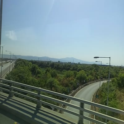 The image shows a view from a bridge overlooking a highway and a landscape with trees and mountains in the distance. On the left, a multi-lane highway with traffic can be seen. Streetlights line the highway. The right side of the image features a curving road with guardrails and vegetation. The sky is clear and light blue. The location is identified as Municipal Unit of Echedoros, Greece, based on the visual cues.