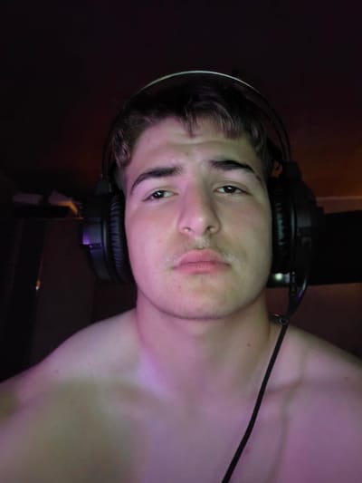 A head and shoulder portrait of a young man wearing black headphones. The man has light brown hair styled with bangs, and a faint mustache. He is looking directly at the camera. The background is dark and out of focus, with hints of purple and pink lighting on the man's shoulders and neck. The image appears to be taken indoors. There is no information in the image to suggest the location is Aleksandrovo, Bulgaria.