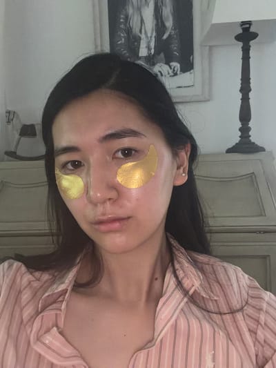 A woman with long, black hair is wearing gold eye masks and a pink and white striped pajama top. She has a fair complexion. The background includes a white cabinet and a framed black and white picture on the wall. This picture does not contain verifiable indicators that confirm the location of Grimaud, France. The scene does not allow verification that this image has been taken in Grimaud, France.