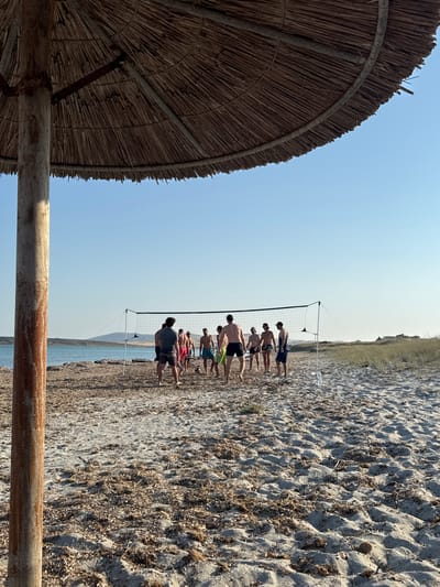 A group of people are playing volleyball on a sandy beach in Moudros Municipal Unit, Greece. The sun is shining brightly in a clear blue sky, suggesting a warm, sunny day. The beach is covered in sand and small rocks, with some dried seaweed scattered about. In the foreground, a large thatched umbrella provides shade. A volleyball net is set up on the beach, and several young men are actively engaged in a game of volleyball. Some are in mid-action, while others are observing or waiting for their turn. The overall atmosphere appears to be one of recreation and enjoyment, with the participants seeming energetic and focused on the game. The water of the sea is visible in the background, calm and reflecting the bright sky. The landscape beyond the beach is arid and covered with vegetation. No visible text is present in the image.