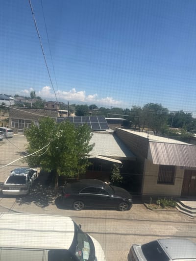 The image depicts a residential area in Metsamor, Armenia, on a clear, sunny day. The foreground shows the tops of several vehicles, including a white van. Moving further back, a black Mercedes-Benz sedan is parked on a paved surface next to a white car. In the middle ground, a large green tree stands prominently, casting shadows on the ground. Behind the tree, a building with a corrugated metal roof is visible, and further back, a building with a tiled roof and stone walls. The background reveals more buildings, some with solar panels installed on their roofs, suggesting a focus on renewable energy in the area. The overall scene is one of a typical urban or semi-urban environment in Armenia, with a mix of residential buildings, vehicles, and natural elements. The sky is bright blue with a few scattered white clouds.
