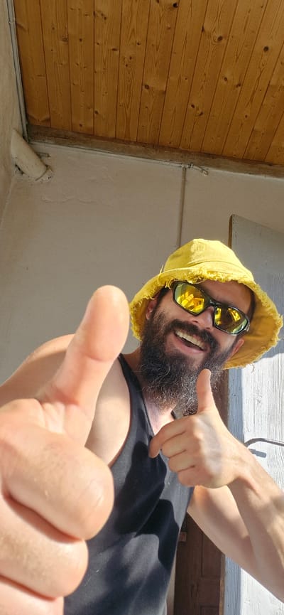 A close-up, low-angle, outdoor selfie shows a man with a beard and a yellow bucket hat, wearing sunglasses and a black tank top. He is smiling and giving a double thumbs-up gesture. The background consists of a textured light-colored wall and a wooden ceiling made of planks. The image appears to have been taken during daylight, with strong shadows indicating sunlight. The scene is consistent with a residential outdoor area, potentially a porch or balcony, in the region of Wattenwil, Switzerland.