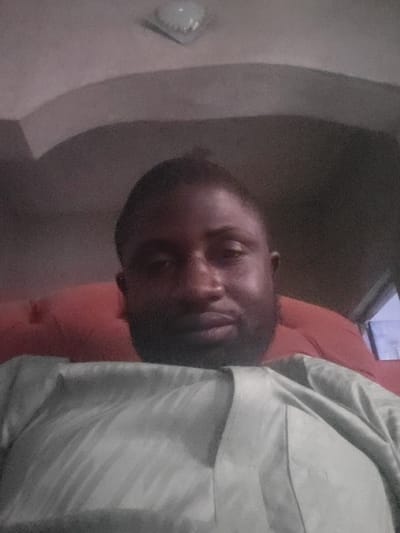 A dimly lit, indoor selfie shows a man with a close-cropped haircut and dark beard wearing a collared shirt. He reclines on a reddish-orange surface. His face is partially obscured by shadows. A blurred window is visible at the bottom of the frame. The background features a curved, white wall with an overhead light fixture. The image is rotated to the left.