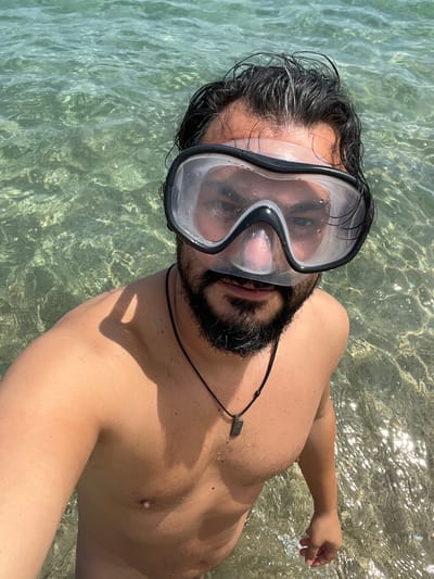 A man with dark hair and a beard, wearing a black and clear snorkeling mask and a black pendant necklace, is standing in shallow, clear, sunlit ocean water. He is looking directly at the camera, taking a selfie. The water is shallow enough to reveal the sandy and rocky seabed beneath. The scene appears to be a sunny day at a beach in Sant Pol de Mar, Spain, likely a tourist area due to the clear water and visible seabed.