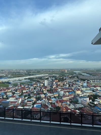 This media file shows an aerial view of a densely populated urban area in Khan Mean Chey, Cambodia. The scene is a sprawling cityscape characterized by numerous buildings with a variety of colored roofs, including red, blue, and yellow. The architecture appears to be a mix of residential and commercial structures, with many appearing to be multi-story buildings. Greenery is interspersed throughout the urban fabric, suggesting parks or residential gardens.

In the foreground, a dark-colored railing with a grid pattern separates the viewpoint from the cityscape below. The railing suggests that the vantage point is from a balcony, rooftop, or observation deck of a tall building. The ground beneath the railing is a dark, textured surface, possibly paving stones or concrete.

The sky is visible on the left side of the image and is a pale blue with scattered clouds. The lighting suggests it is daytime, likely late morning or early afternoon. The overall atmosphere appears calm and clear, with no immediate signs of intense weather. The orientation of the image is vertical, with the cityscape extending upwards from the bottom of the frame and the sky occupying the upper left portion. No people are clearly visible in the scene, and there is no discernible text in the image.