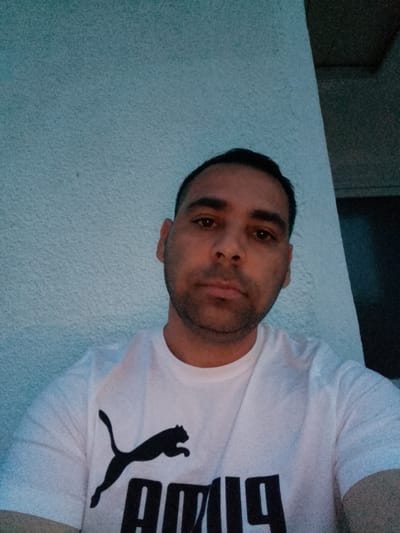 The image is a selfie taken outdoors, featuring a man with medium skin tone, dark hair, and a beard. He is wearing a white t-shirt with a black Puma logo and text. The background consists of a textured blue wall and a darker structure, possibly a building or wall, to the right. The lighting suggests it might be evening or dusk. The specific location in Bukovlak, Bulgaria cannot be determined from the visual information provided in the image.