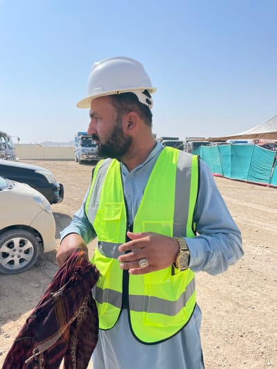 In Pir Dust, Afghanistan, an adult male stands outdoors under a clear blue sky. He is depicted from the chest up, in profile, facing right. He wears a white hard hat, a neon yellow high-visibility safety vest with grey reflective stripes over a light blue long-sleeved traditional garment, and has a dark beard and mustache. His left hand holds a folded, patterned fabric, exhibiting red, brown, and black tones. His right hand is open, fingers slightly splayed, mid-gesture. A gold-colored wristwatch is on his left wrist, and a large ring with a light-colored oval stone is on his right hand. The ground consists of dry, unpaved dirt. In the background, several vehicles are present, including a light-colored compact car, a dark car, and multiple blue and white heavy-duty trucks. To the right, green tarpaulin structures and a portion of a white tent are visible. A light-colored wall and distant, low hills complete the horizon.